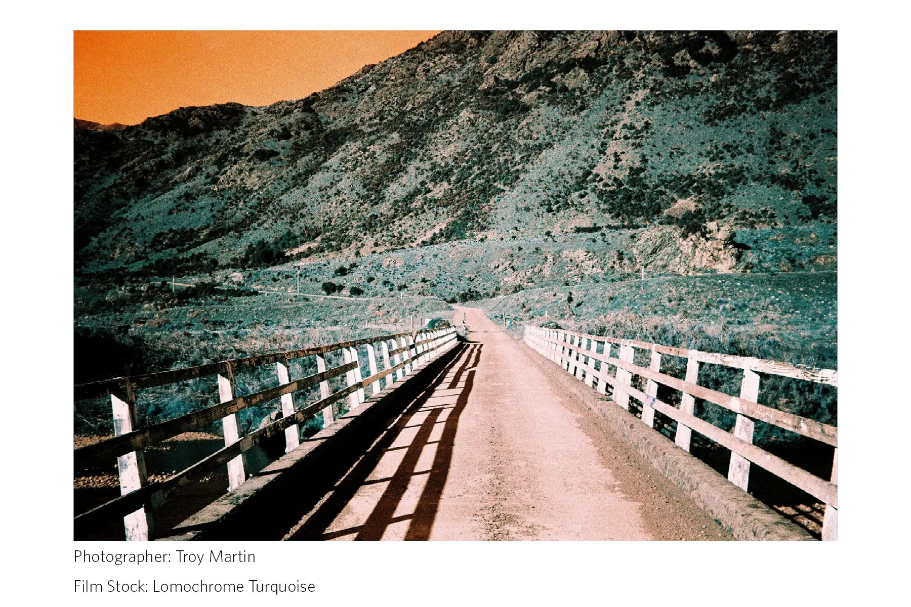 Winding road through a mountainous landscape with a white fence on either side, shot with Lomochrome Turquoise film.